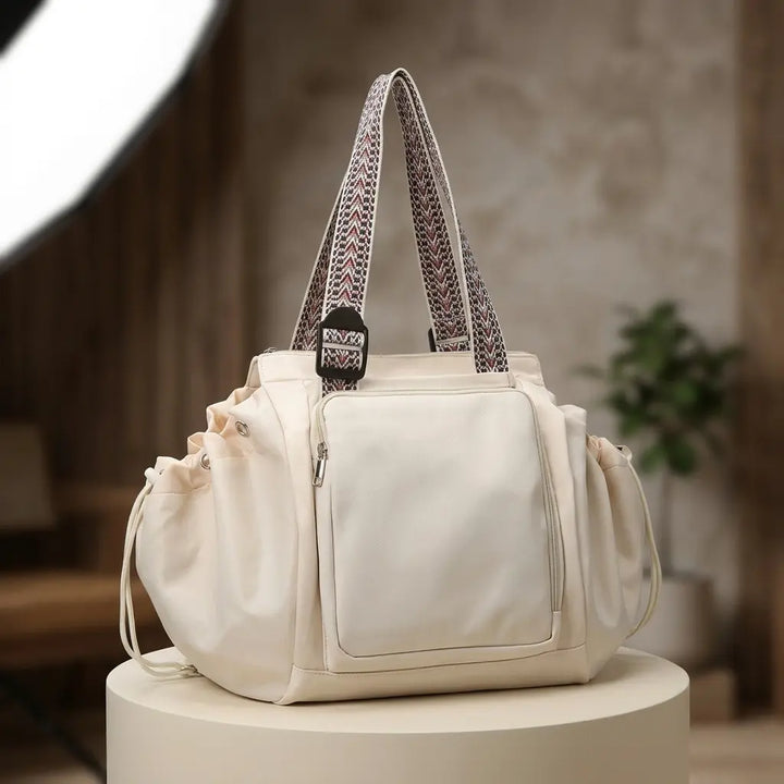 Cream convertible tote with front zip pocket and drawstring sides on a pedestal
