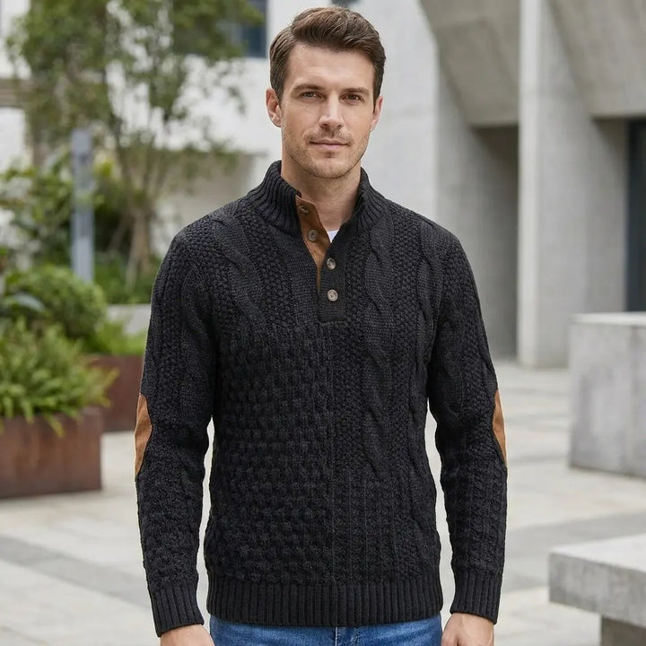 Men's cable knit sweater in black with elbow patches in outdoor setting, front model view